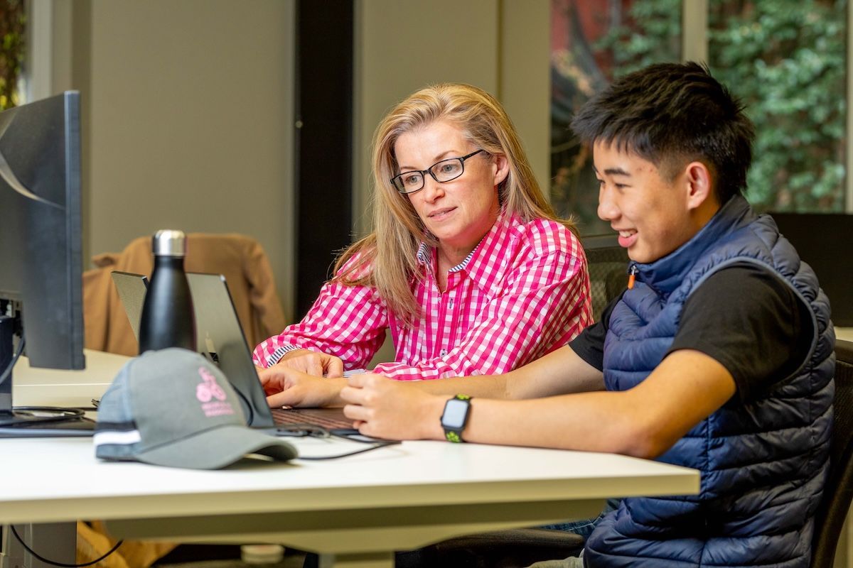 Woman in pink shirt and Asian man at a laptop, smiling. They sit at a white table in a bright room.