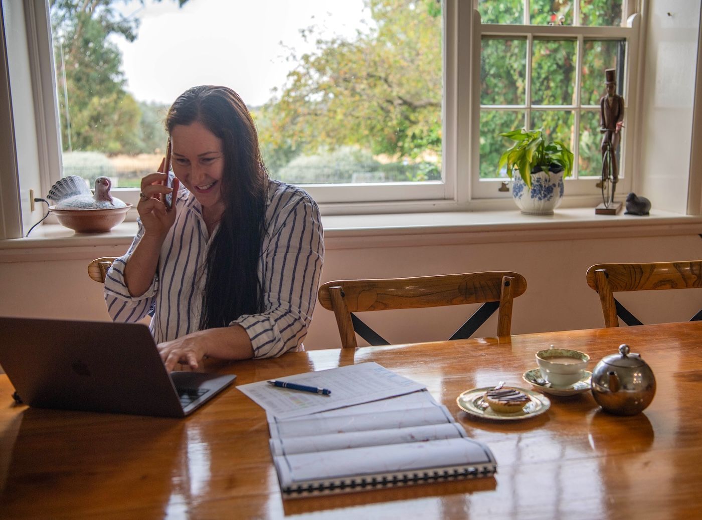 Woman smiling, on phone, working on laptop at a table. Papers, tea, and window view.