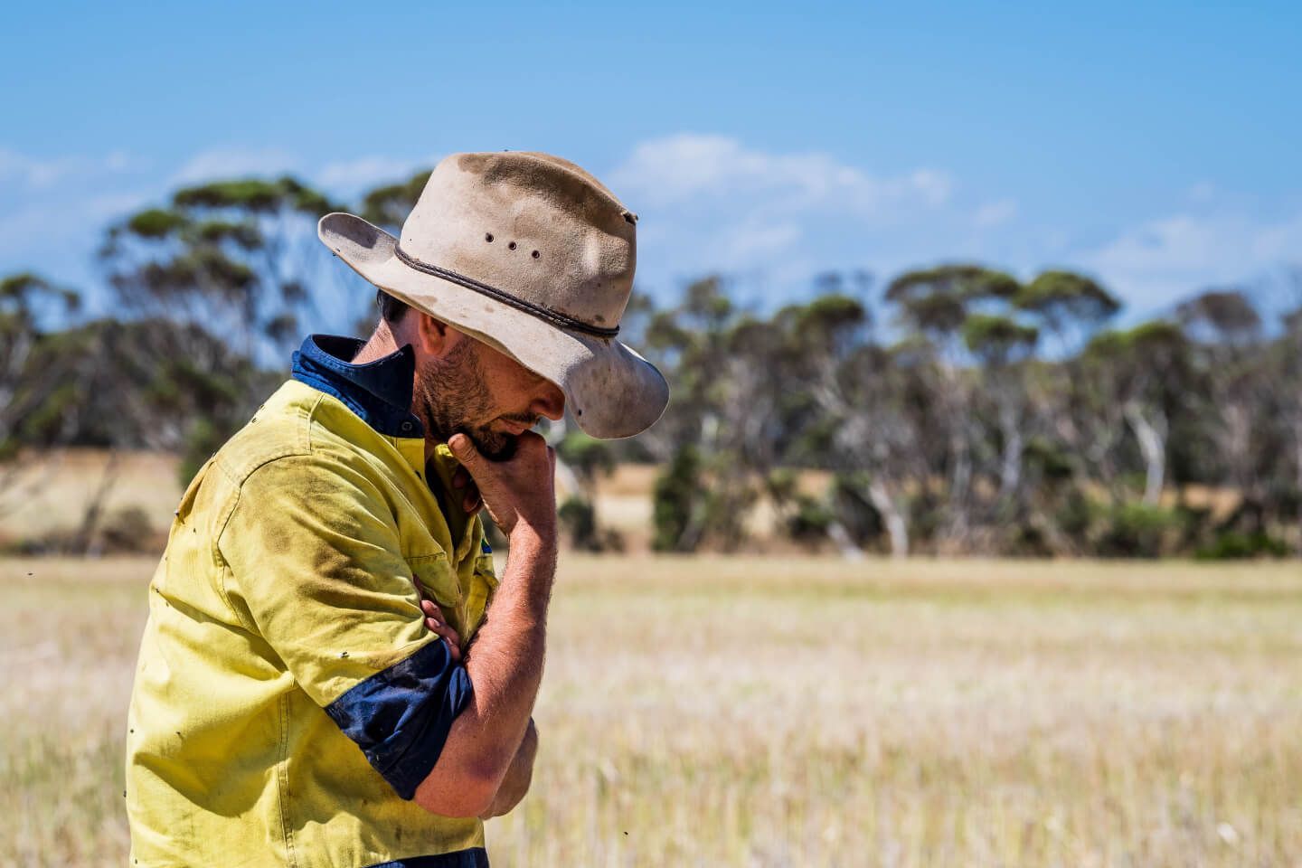 A farmer in a hat and yellow shirt looking down pensively in a dry field under a sunny sky.