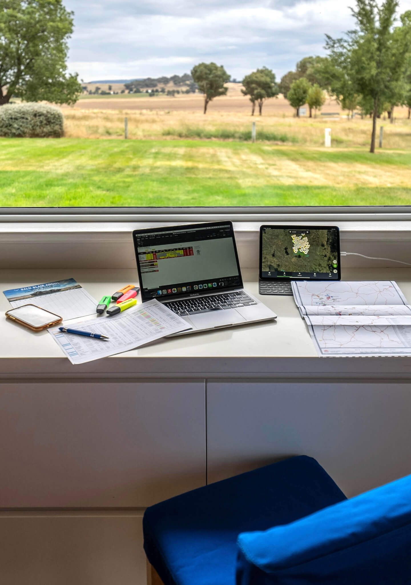 Desk with laptops and papers, overlooking a green field with trees under a cloudy sky.
