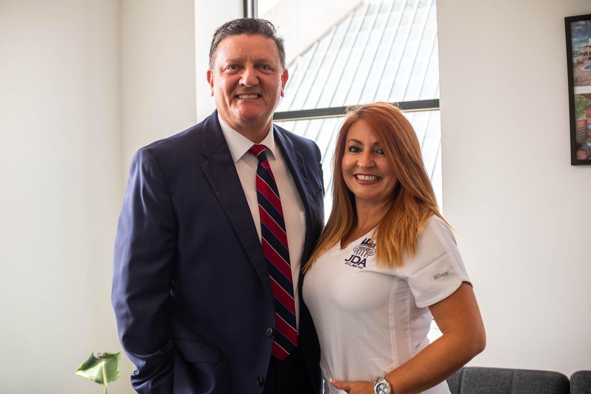 Man in suit and woman in white shirt smile, posing together indoors.