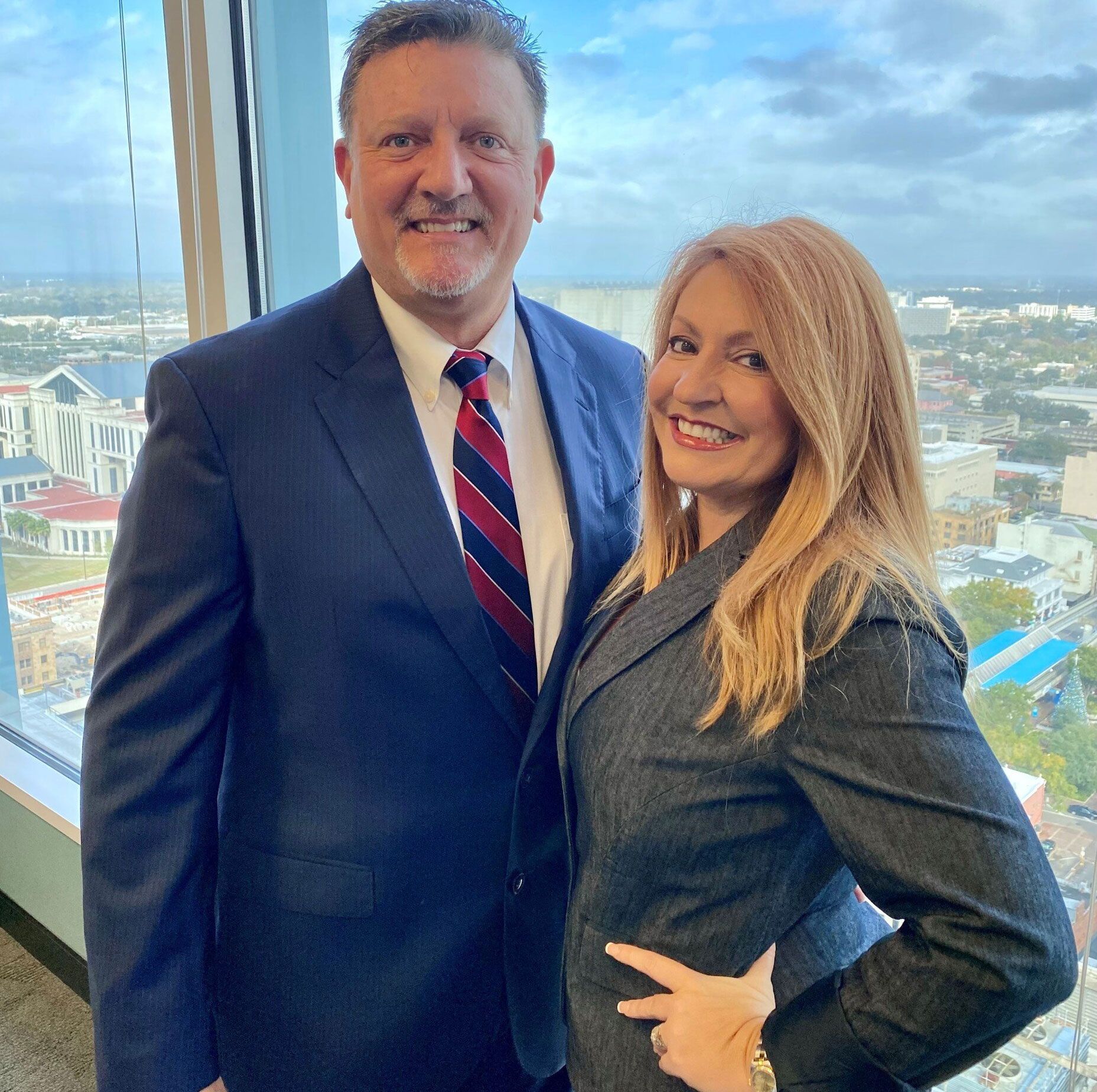 Man and woman in business attire smiling by a large window overlooking a city.