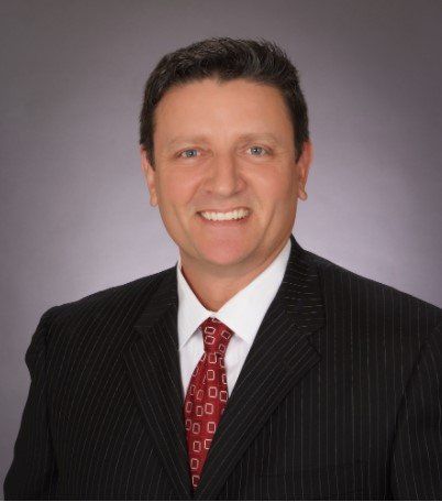 Man in suit, red tie, smiling. Gray background.