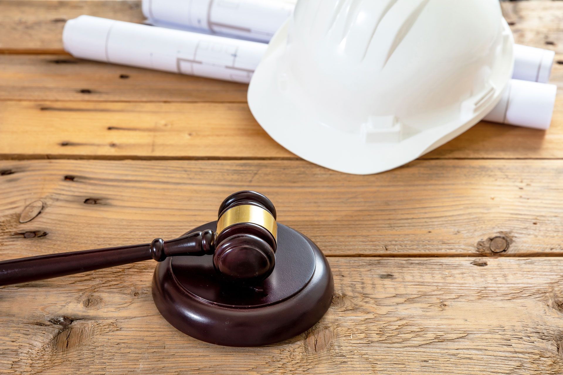 Gavel and sound block next to a white hard hat and rolled-up blueprints on a wooden surface.