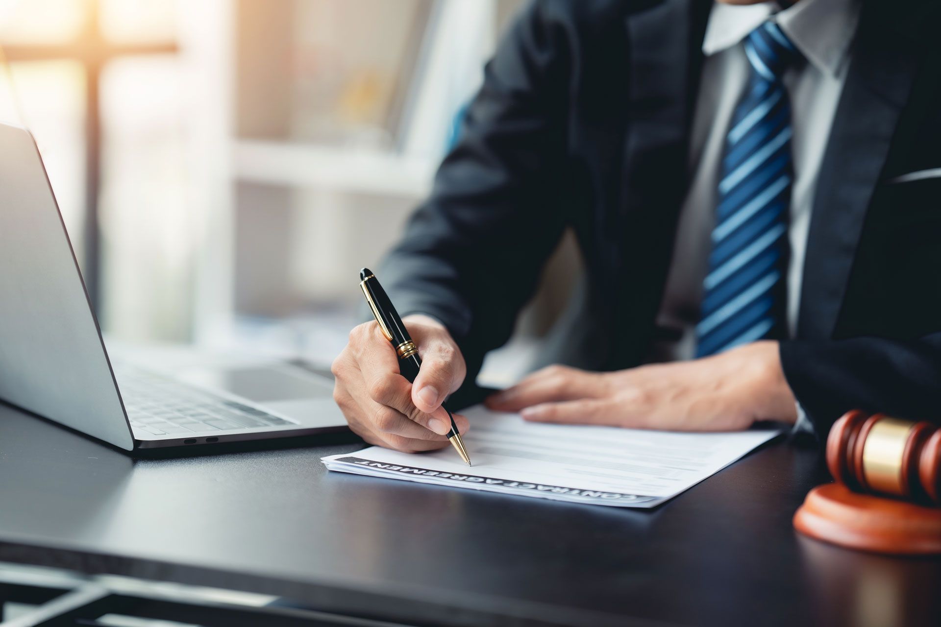 A lawyer in a suit writes on a document at a desk with a laptop and gavel nearby.