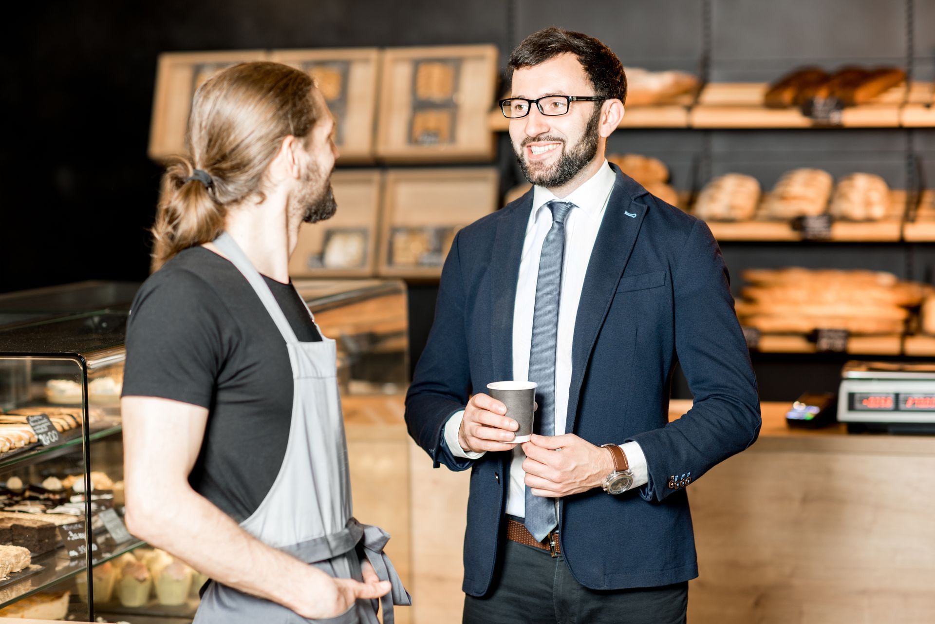 A man in a suit and tie is talking to a man in an apron in a bakery.