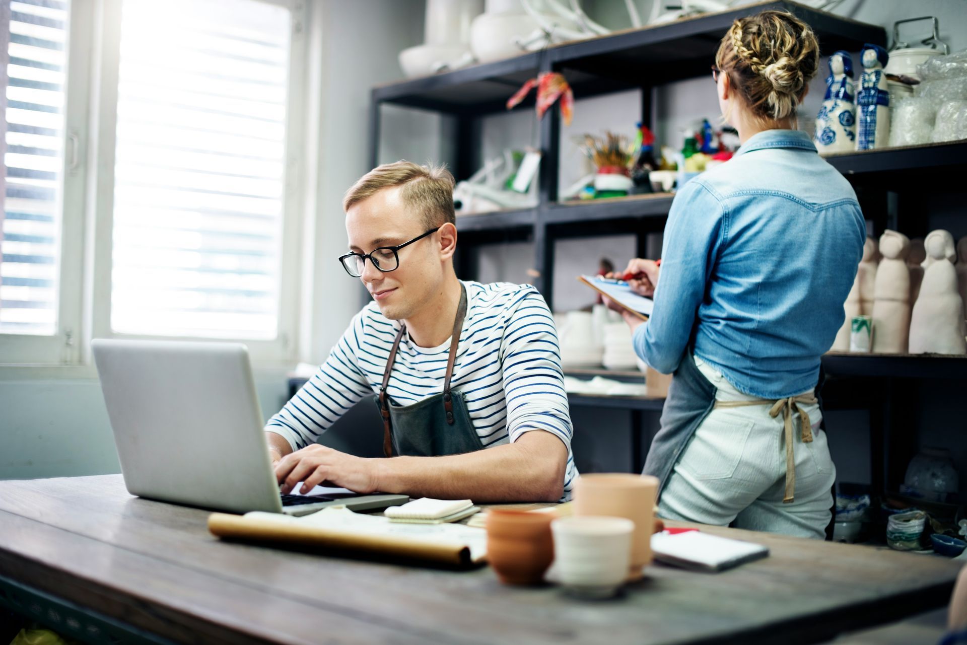 A man is sitting at a table using a laptop computer while a woman stands behind him.