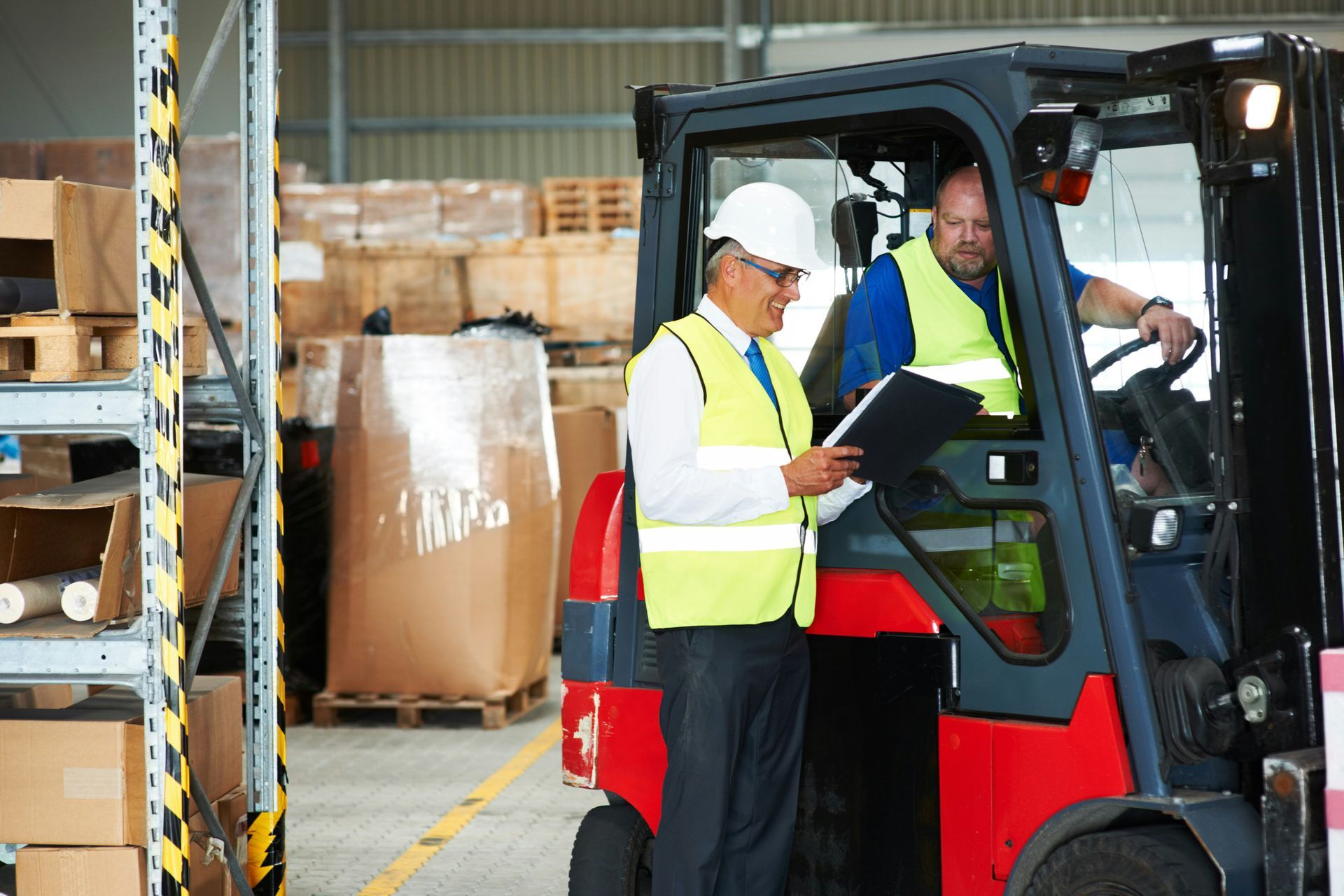 Two men are standing next to a forklift in a warehouse looking at a clipboard.