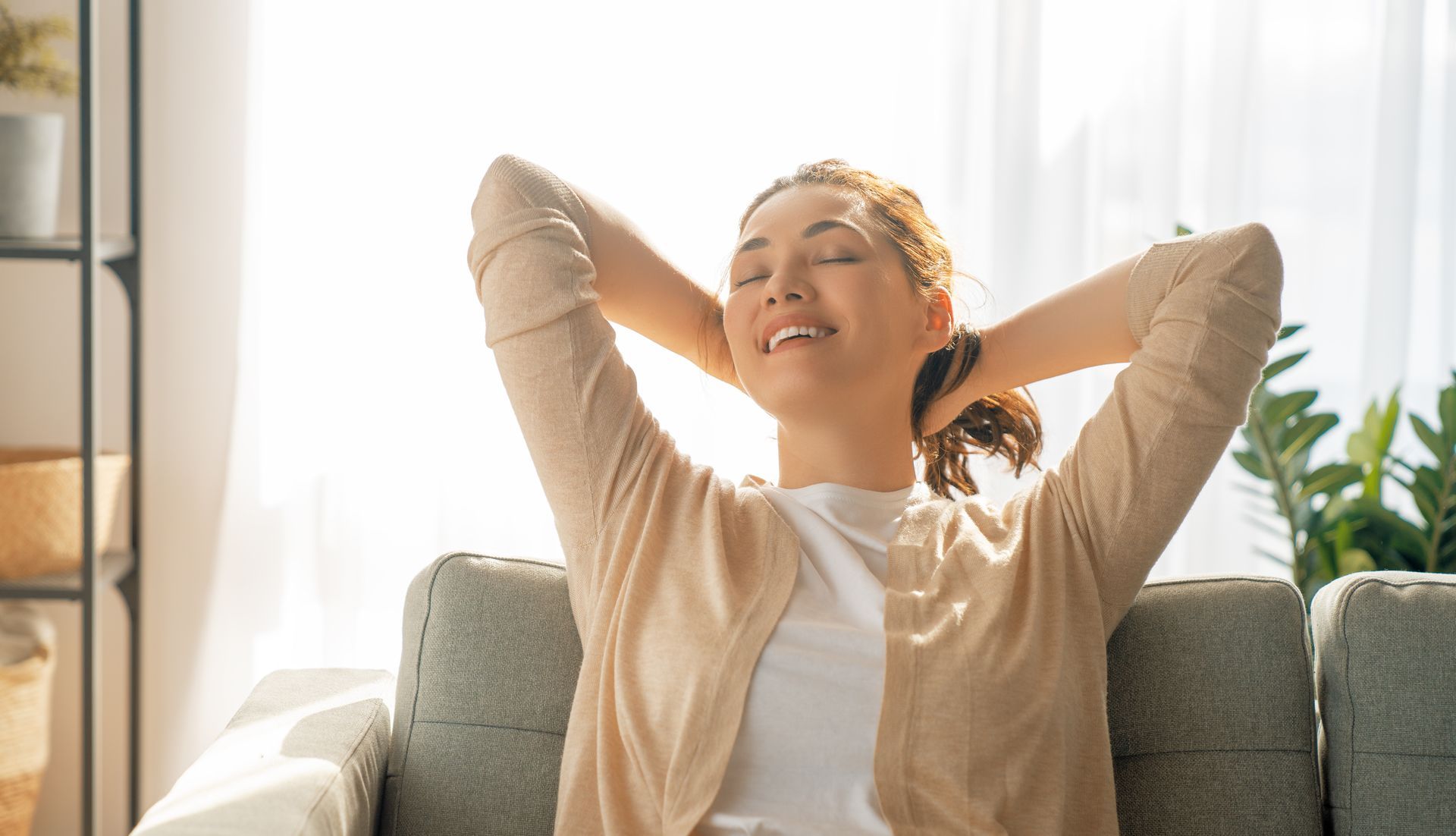 A woman is sitting on a couch with her eyes closed and her hands behind her head.