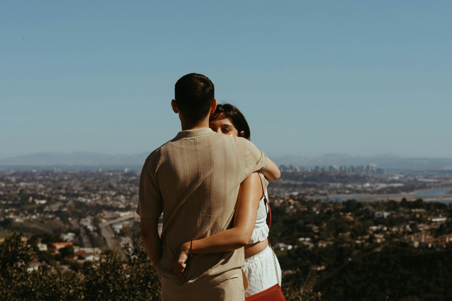 Couple embracing on a hill overlooking a cityscape on a sunny day.