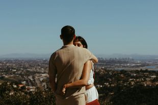 Couple embracing on a hill overlooking a cityscape on a sunny day.