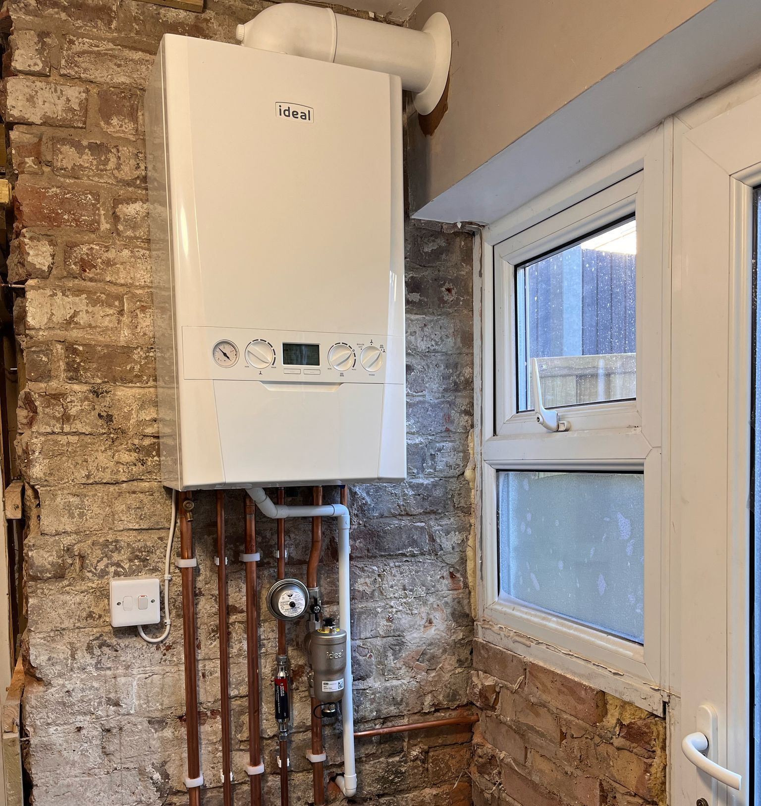 A white boiler is hanging on a brick wall next to a window.