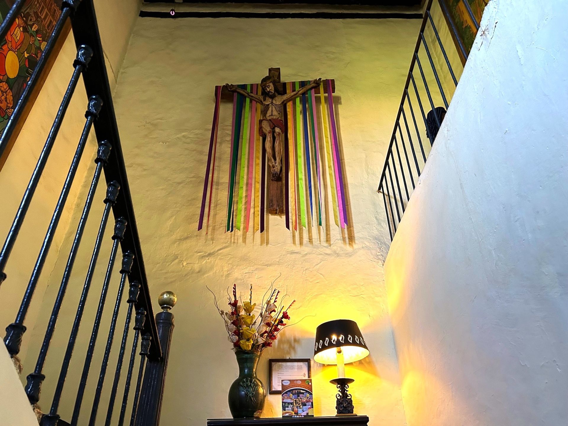 A wooden crucifix hangs on a textured beige wall between a staircase, adorned with colorful ribbons and lit by a lamp.