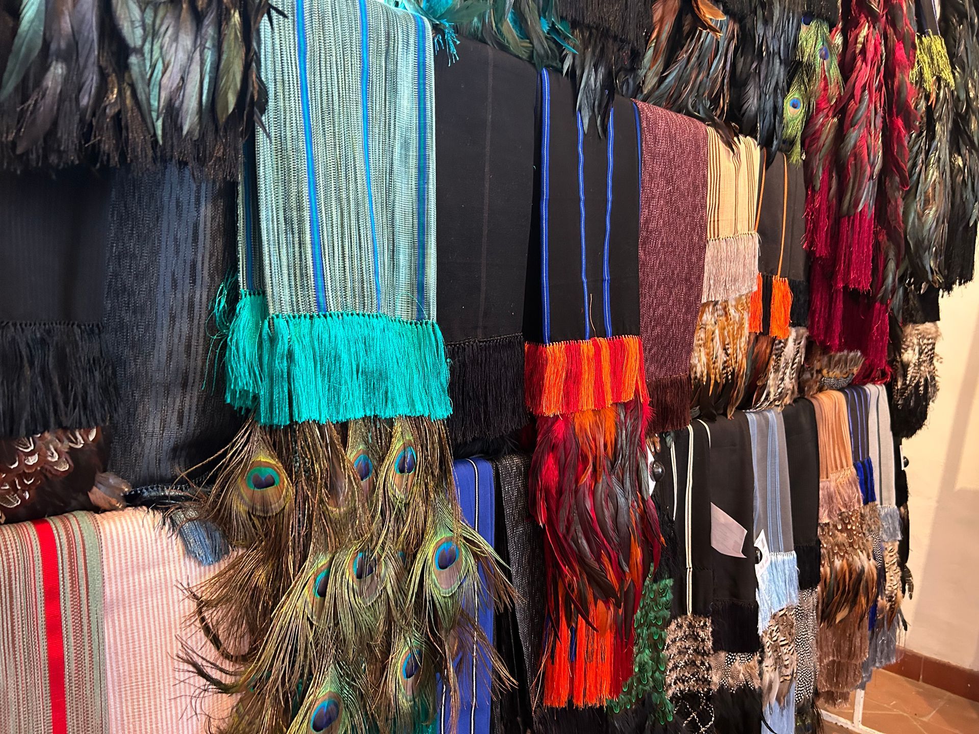 Display of colorful textile scarves with fringed edges and peacock feathers, hanging on a wall.