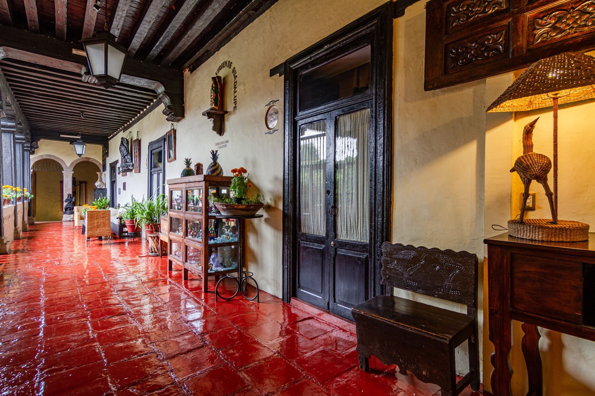 Red-tiled covered walkway with dark wooden beams and trim. Antique furniture, plants, and decorative items line the walls.