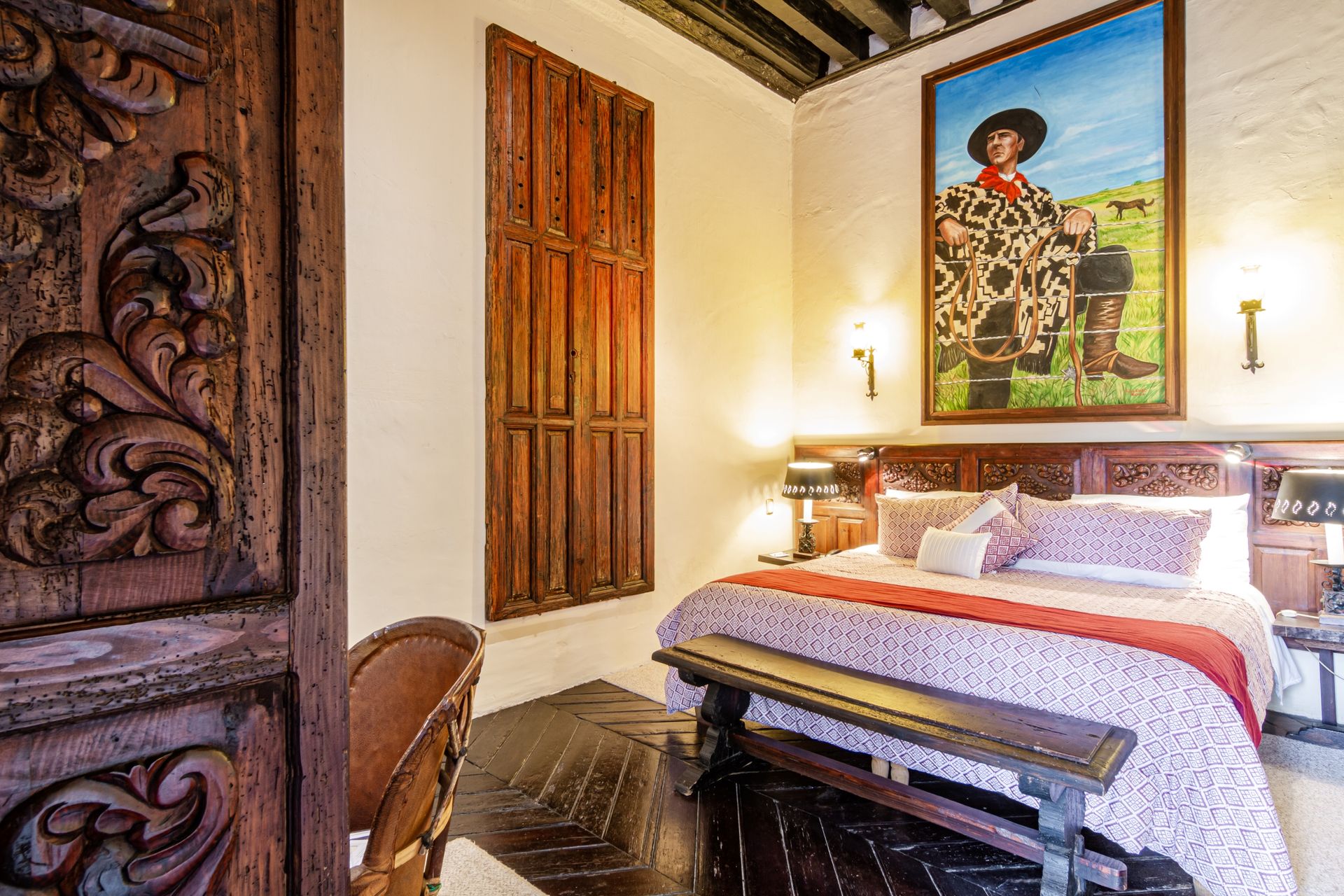 Bedroom with a large bed, artwork, a carved wooden door, and a bench. Warm lighting and a traditional style.