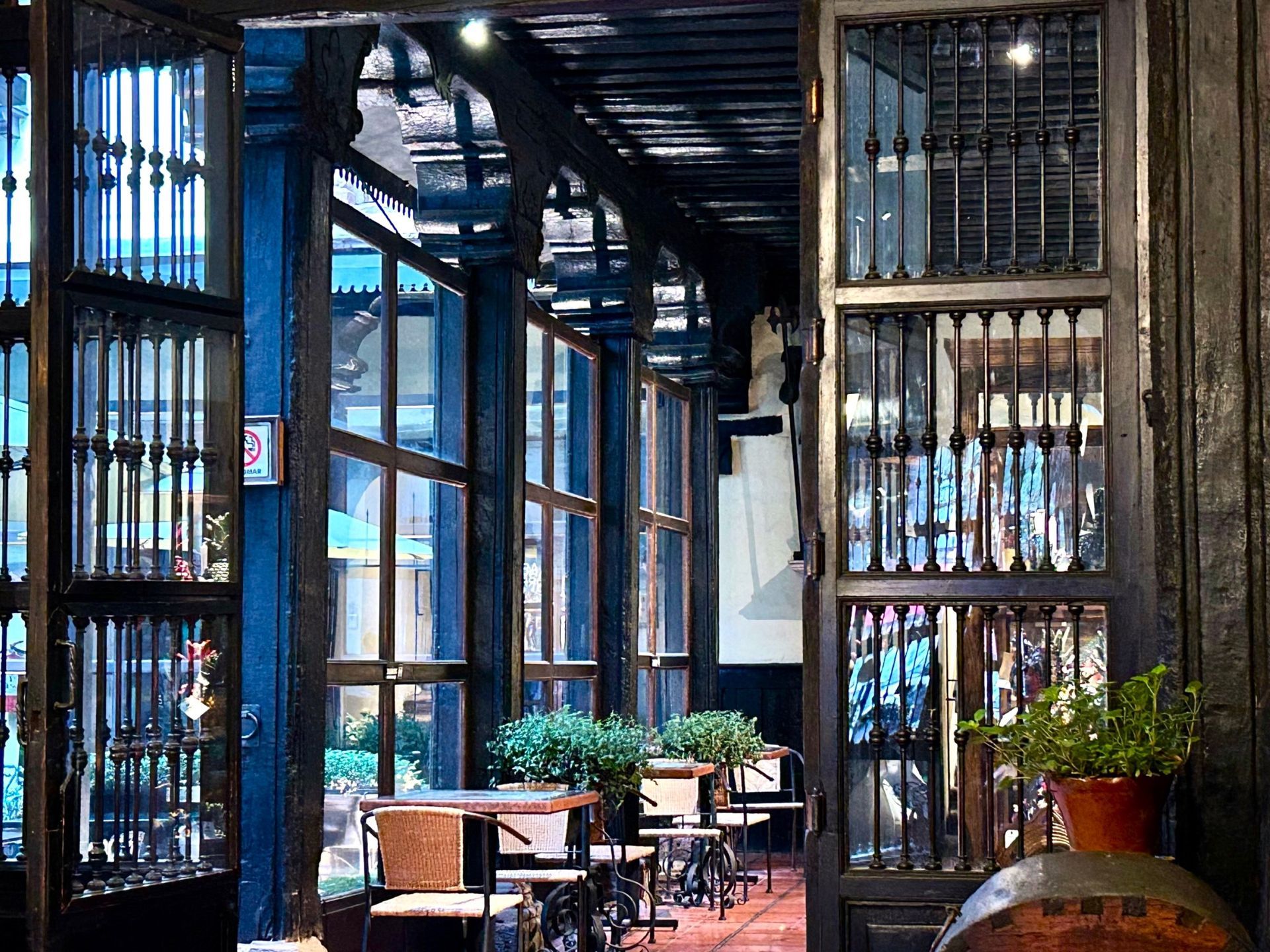 Dark wooden framed windows and door to a cafe with tables and plants inside. Blue sky is visible through the glass.