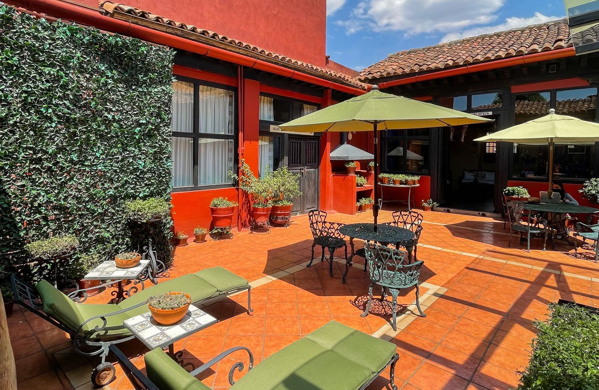 Outdoor courtyard with tables, chairs, and umbrellas. Green lounge chairs sit near a wall of foliage.