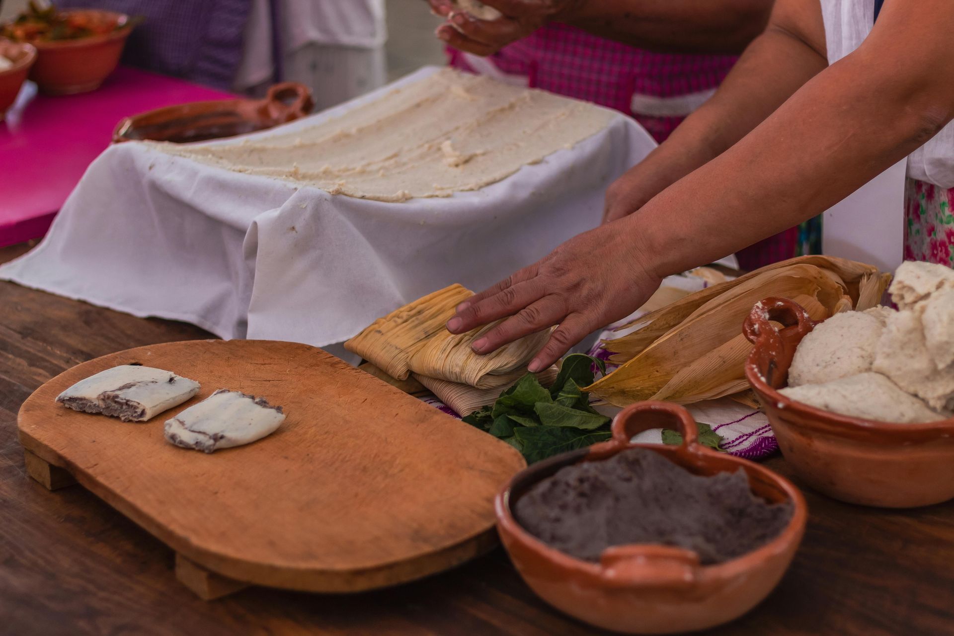 Person preparing Mexican food. Includes tortillas, tamales, beans, and other ingredients on a wooden board and table.