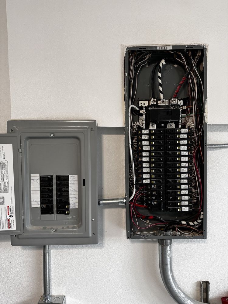 Electrical panel, two gray metal boxes mounted on a white wall, with various wires visible.