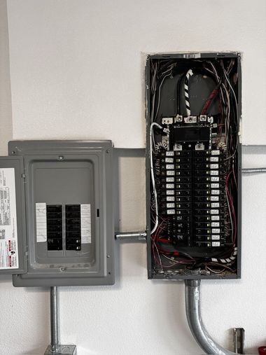 Electrical panel, two gray metal boxes mounted on a white wall, with various wires visible.
