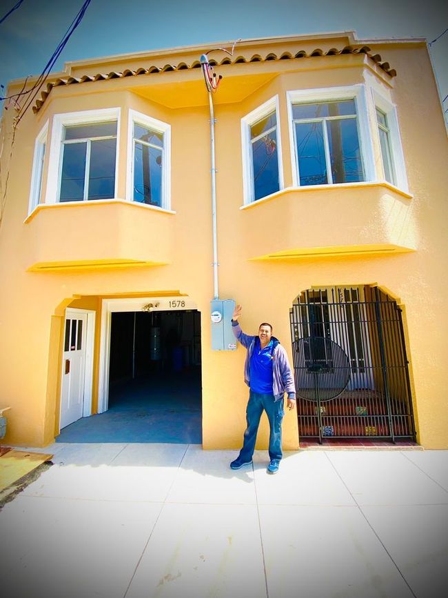 Man stands in front of a yellow house with a garage and windows. He points to an electrical box.