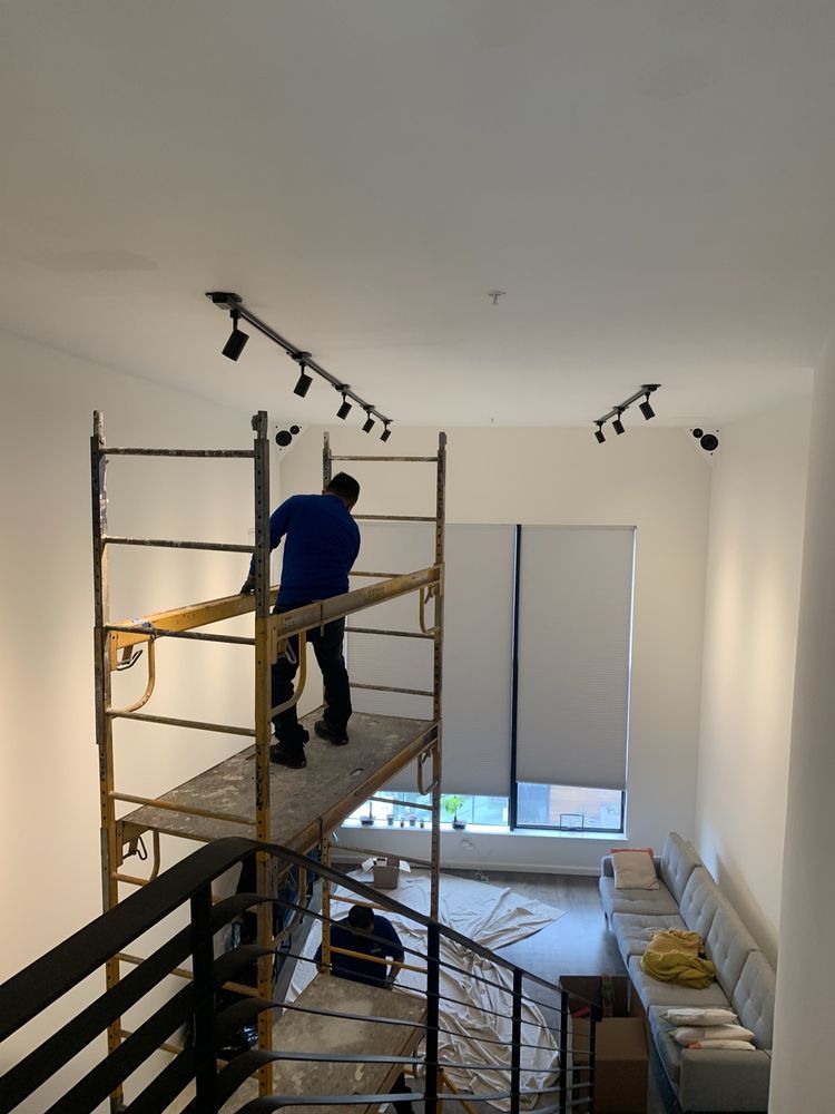 Man on scaffolding installing track lighting in a high-ceilinged room with a staircase.