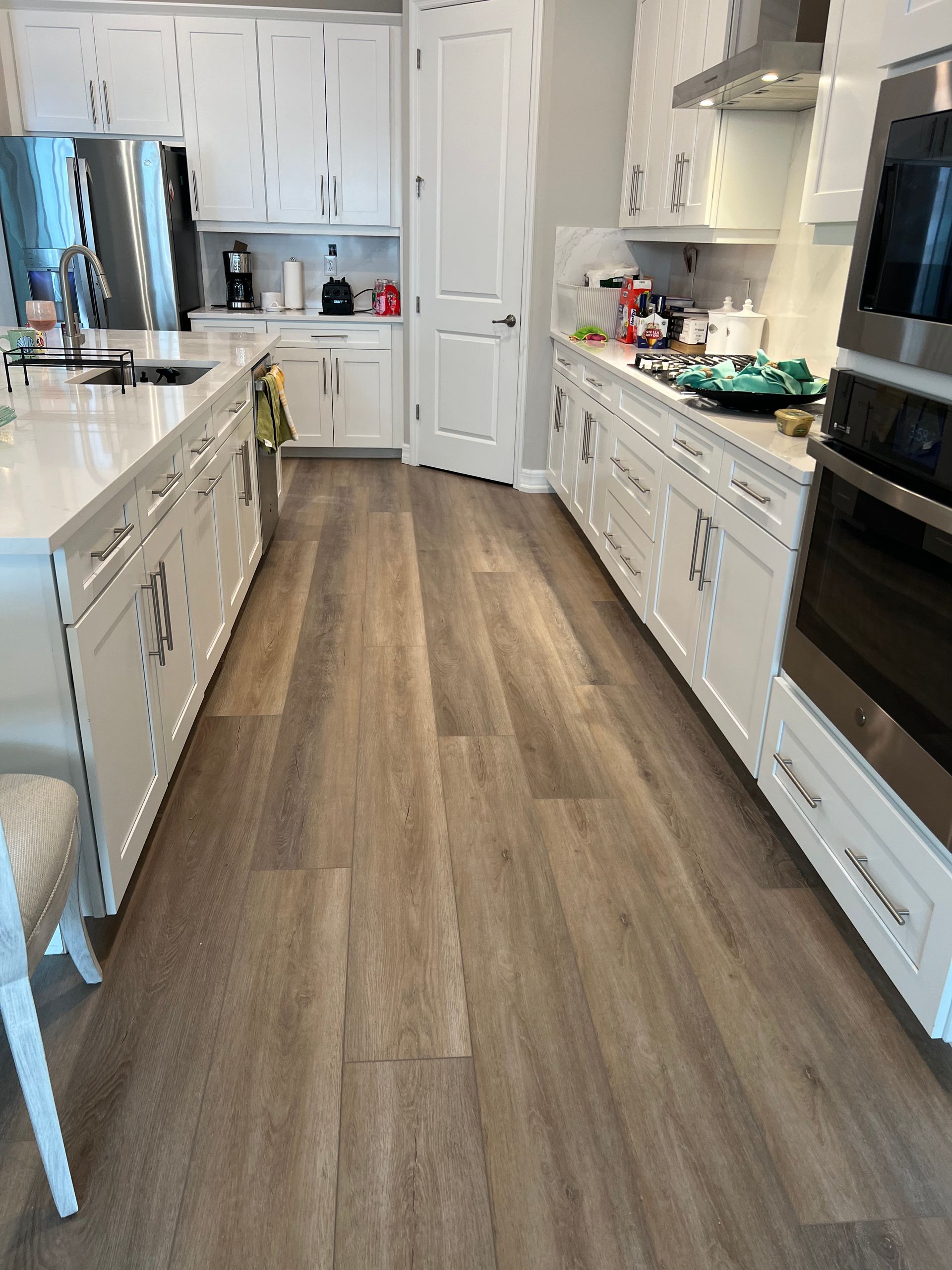 A kitchen with white cabinets, a long island, and wood-look flooring. The kitchen has an oven, stovetop, and a white countertop.