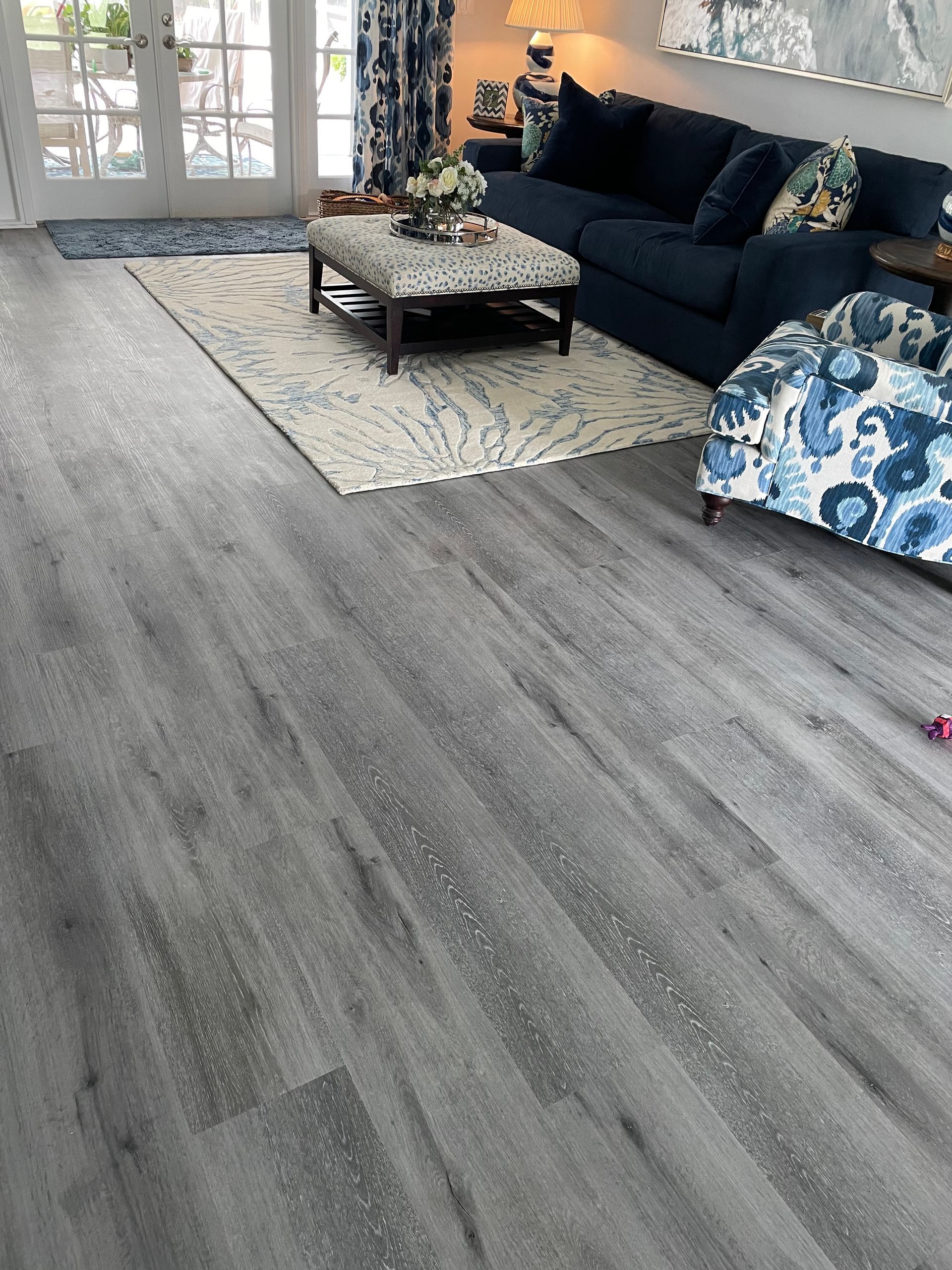 Gray wood-look flooring in a living room with blue furniture, a patterned rug, and glass doors leading to a porch.