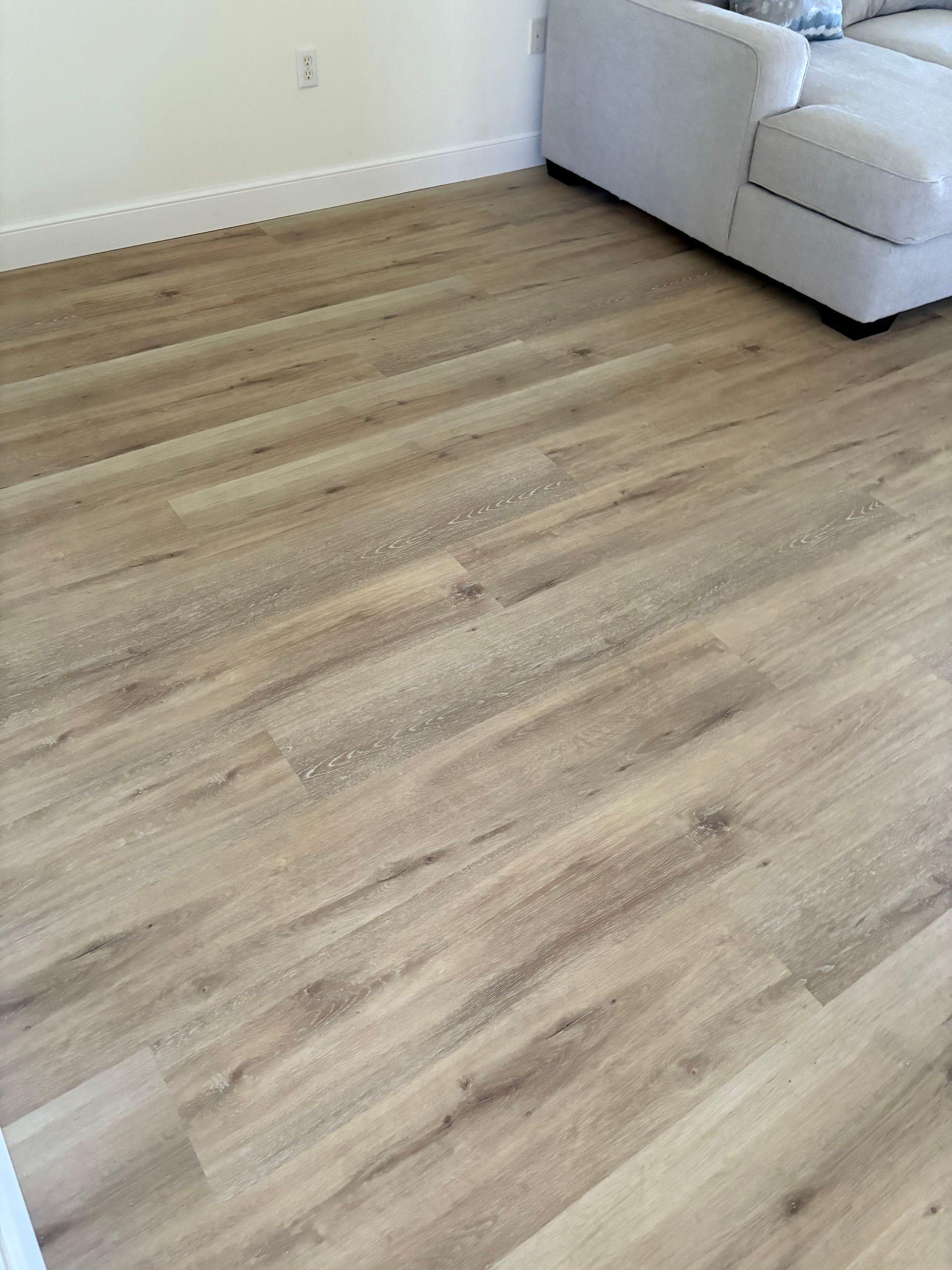 Light brown wood-look flooring in a room, partially visible white sofa in background.