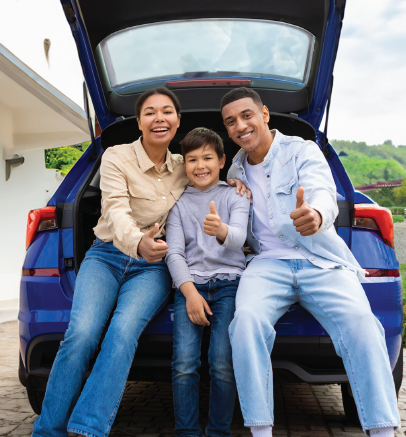 Familia sentada en el maletero de un coche, sonriendo y haciendo un gesto de aprobación. Coche azul, al aire libre.