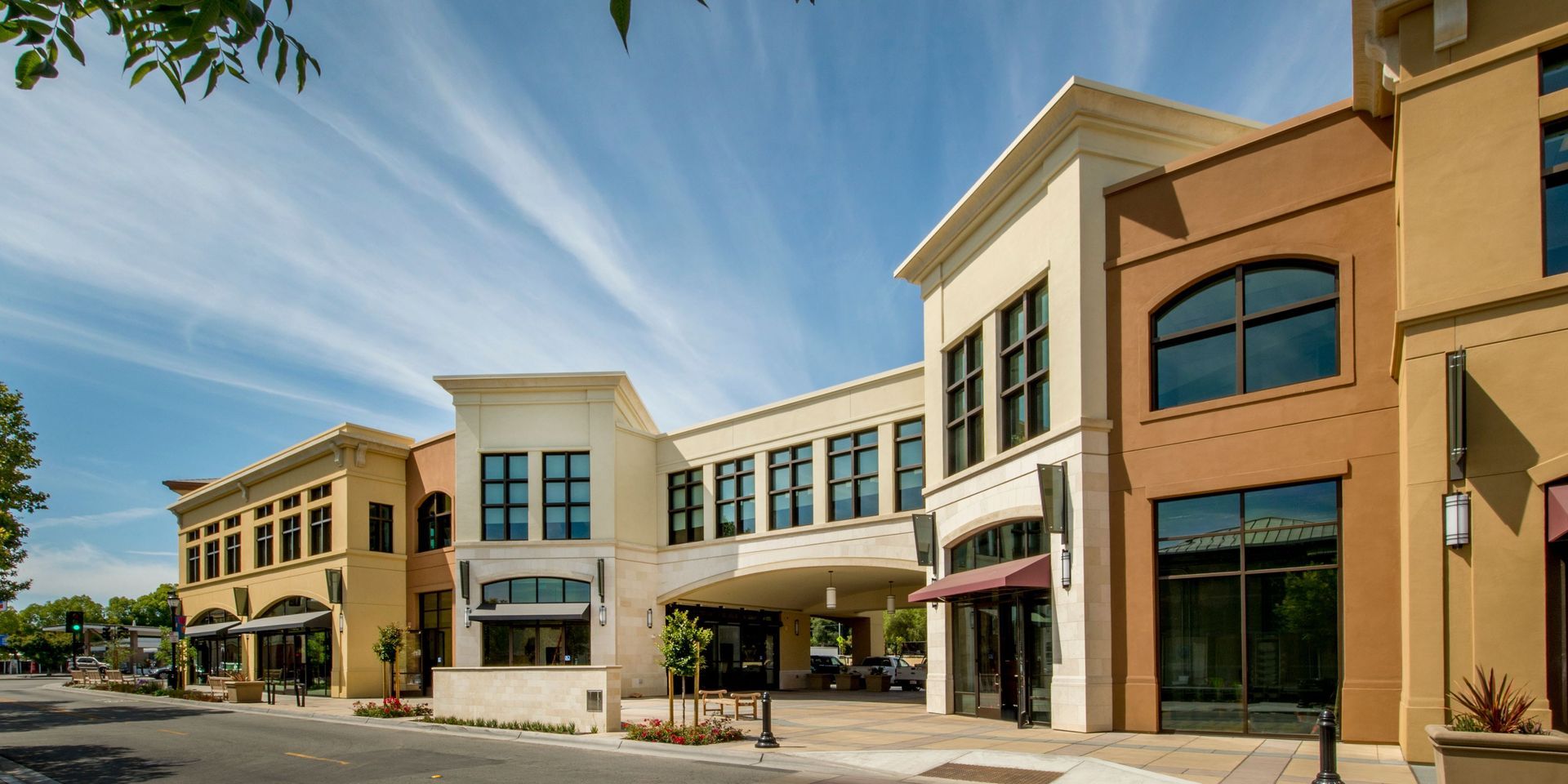 Shops and offices line a street on a sunny day; architecture features beige, brown, and tan buildings under a blue sky.