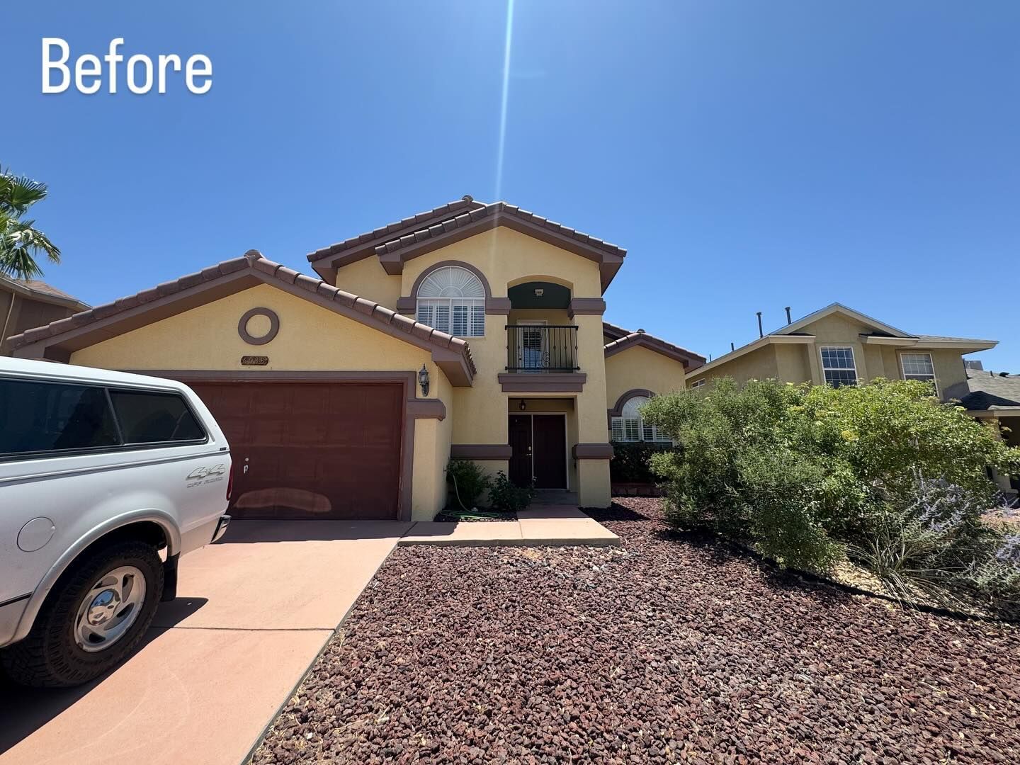 Tan two-story house with red roof, brown garage door, and red gravel landscaping. Bright blue sky.