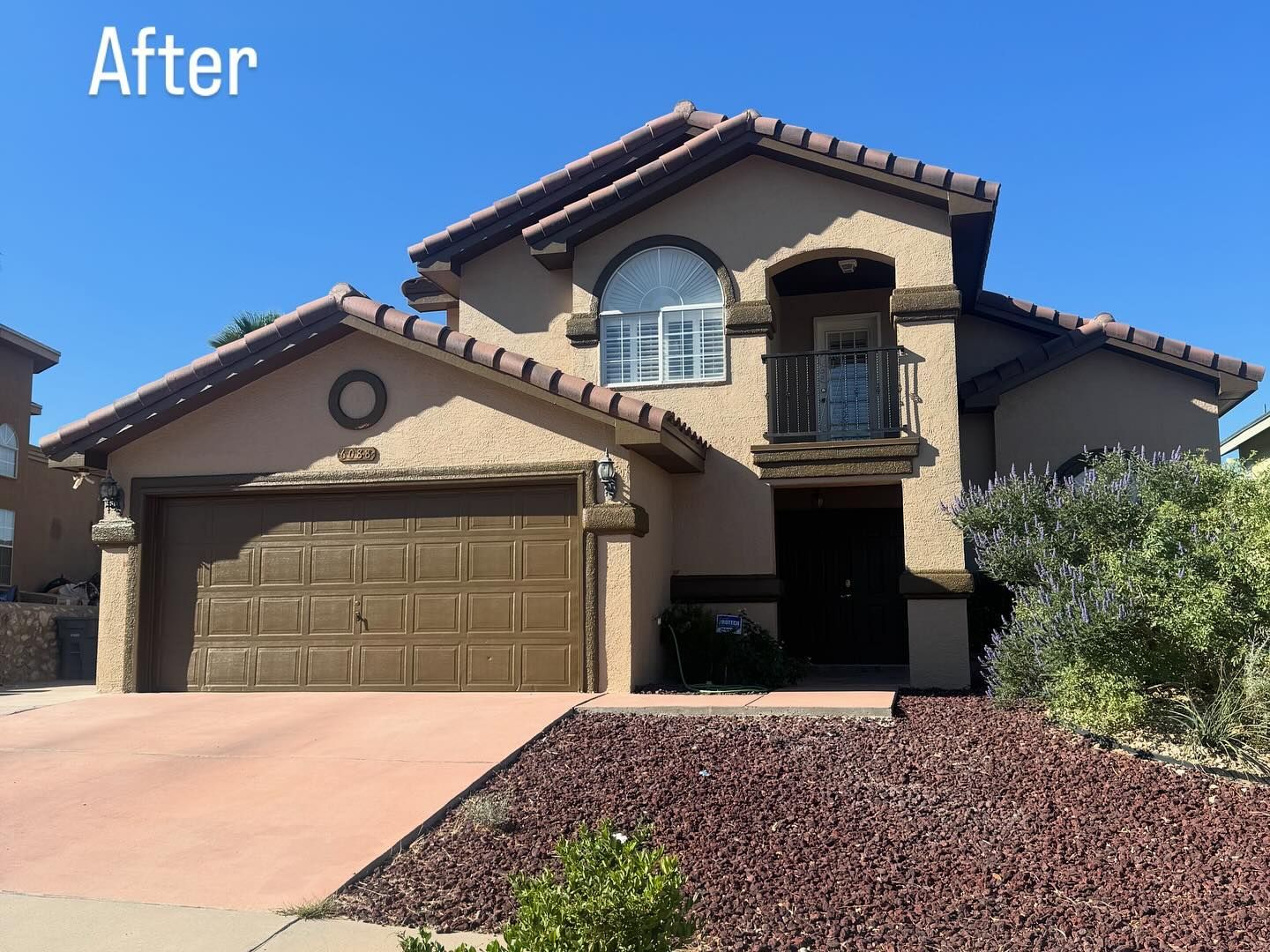Two-story stucco house with brown roof tiles and garage door, red driveway, and a small patch of red mulch.