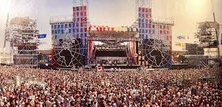 Large crowd at an outdoor music concert; stage with colorful tower structures and global map.