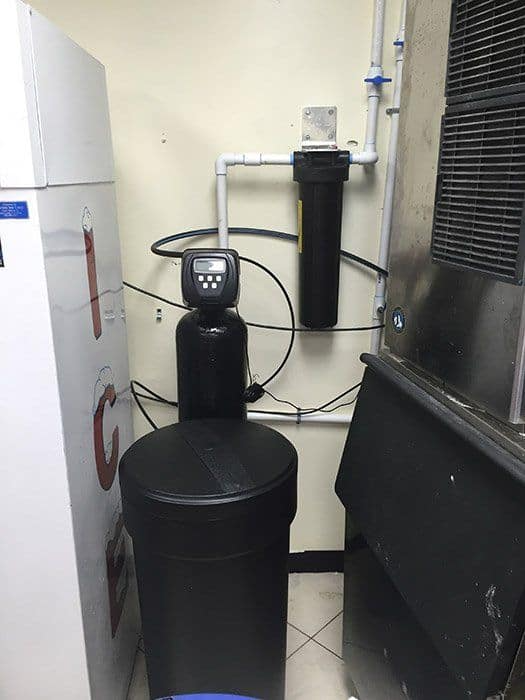 A black water softener is sitting in a room next to a refrigerator.