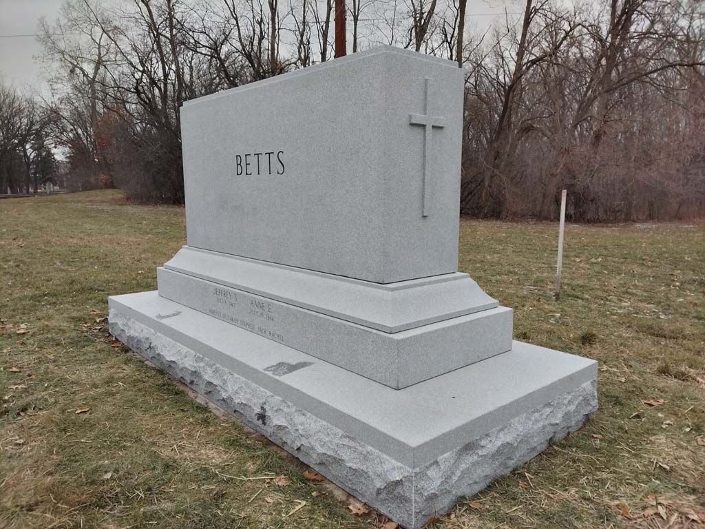Gray stone mausoleum with a cross on the side, standing in a grassy cemetery area.