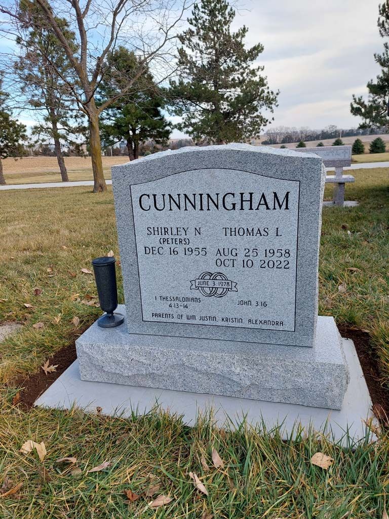 Gray granite Cunningham headstone in a cemetery, with grass, trees, and a cloudy sky behind it.