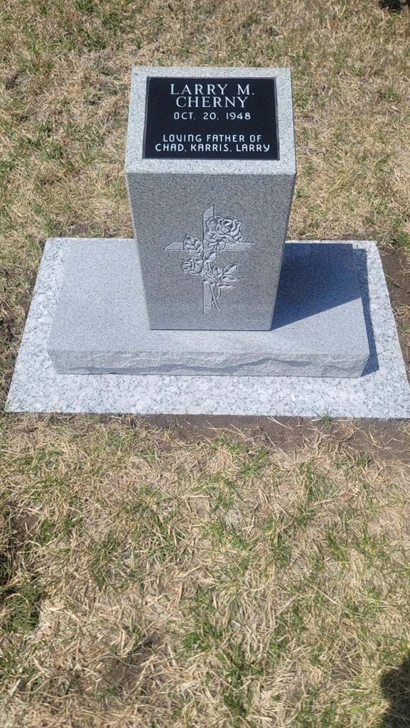 Small gray memorial monument with a black plaque, set on a concrete base in a grassy area.
