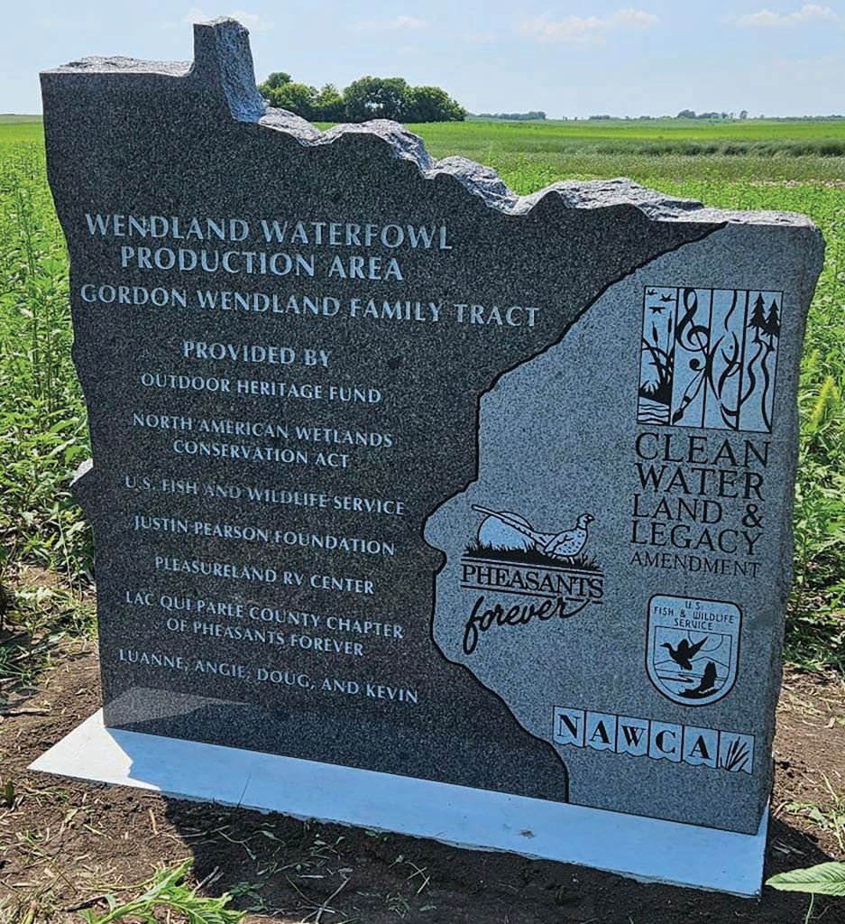 Dark stone memorial sign in a field, with engraved text and logos, honoring a wind and water conservation site