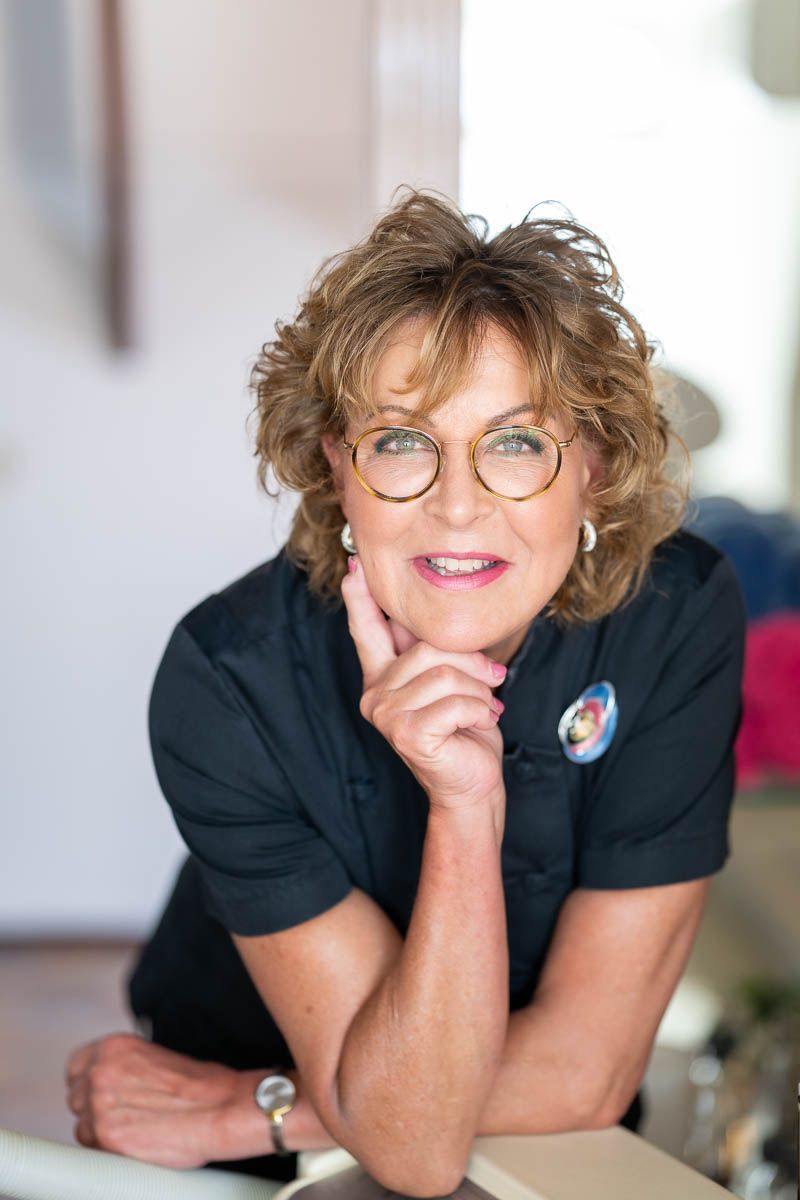 Een vrouw draagt een bril en een zwart shirt en lacht met haar hand op haar kin.