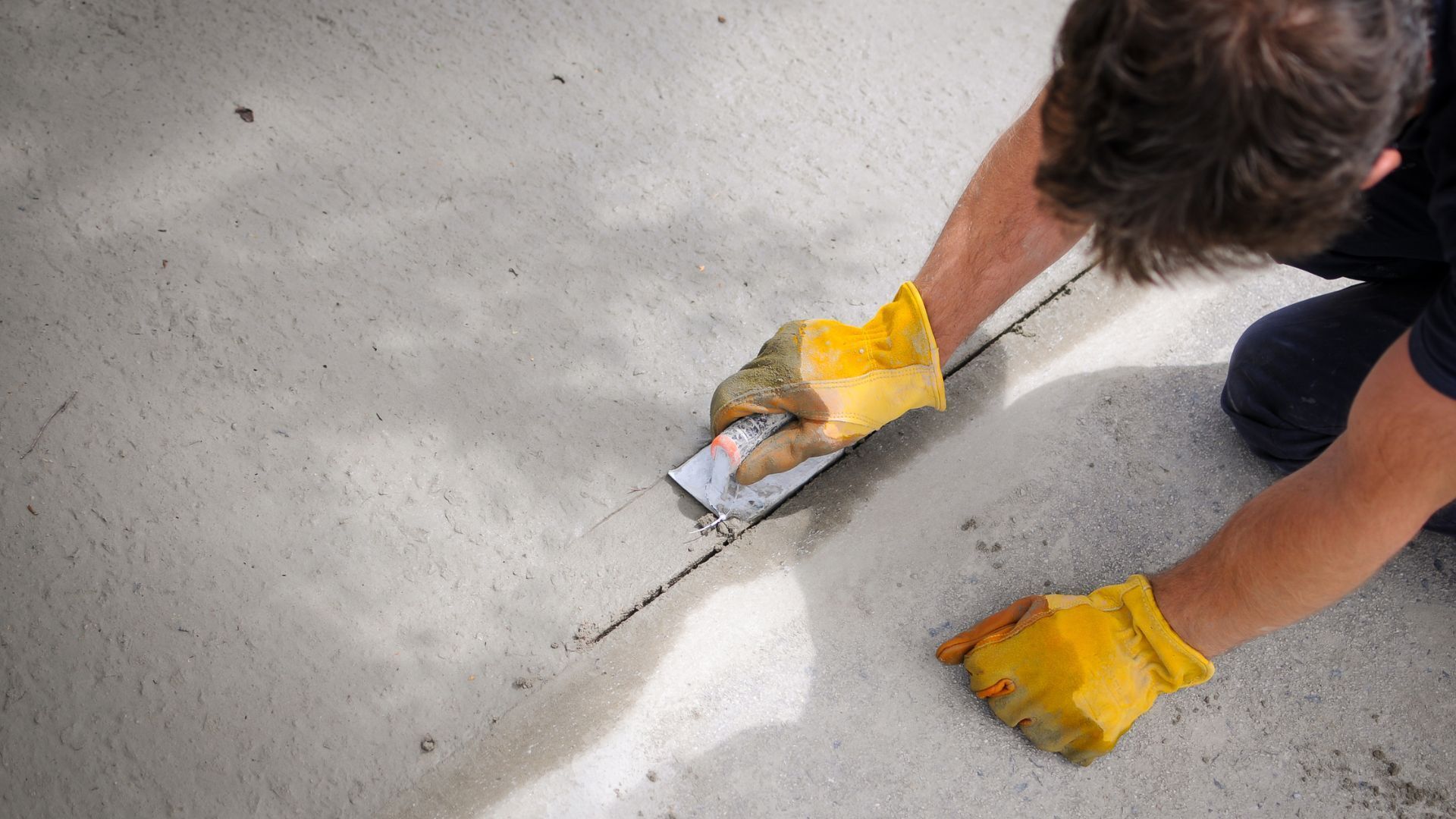 A man wearing yellow gloves is kneeling down and using a trowel to spread concrete.