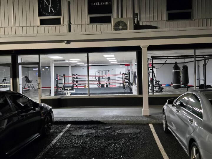 Boxing gym storefront at night, showing ring, equipment, and parked cars.