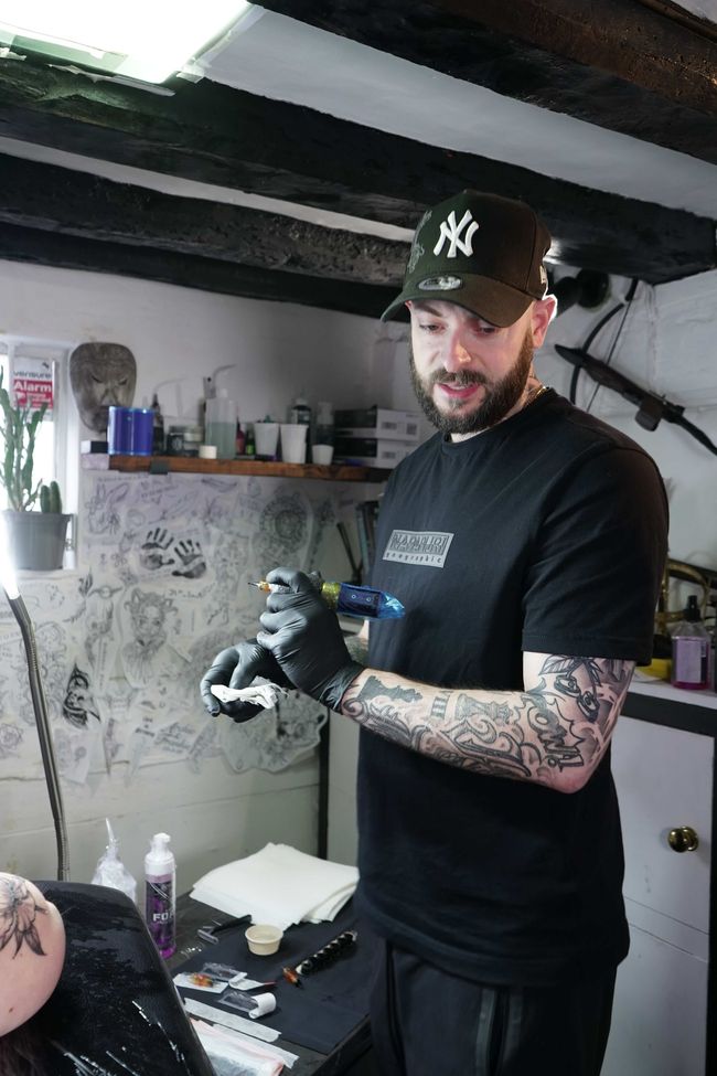 A tattooed artist sits in a dimly lit, industrial-style tattoo studio, with equipment and clients in the background.