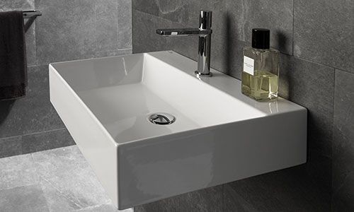 un lavabo a forma quadrata di color bianco con rubinetto in acciaio si intravede uno specchio e un portasciugamani con un asciugamano marrone