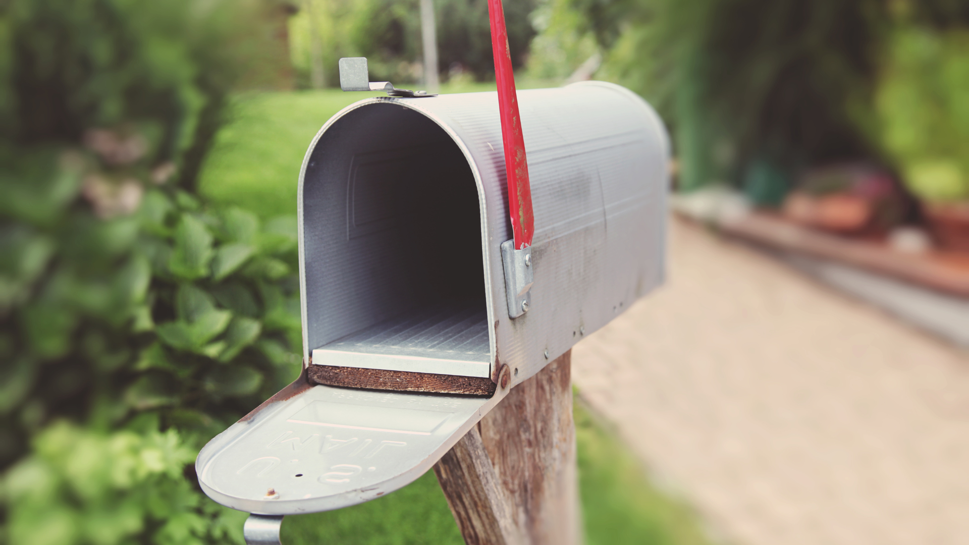Open, empty, silver mailbox with red flag, mounted on a wooden post; blurred background of greenery and path.