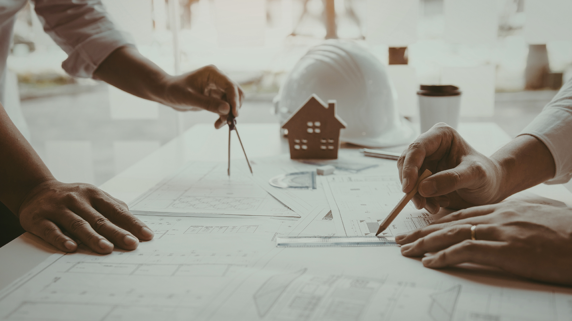 Two people reviewing blueprints with tools and a model house on a desk.
