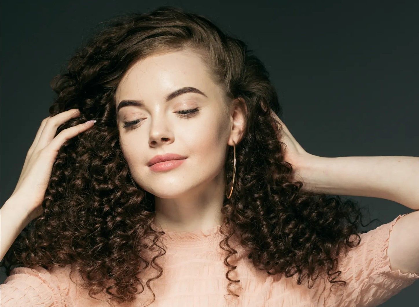 A woman with curly hair is touching her hair with her hands.