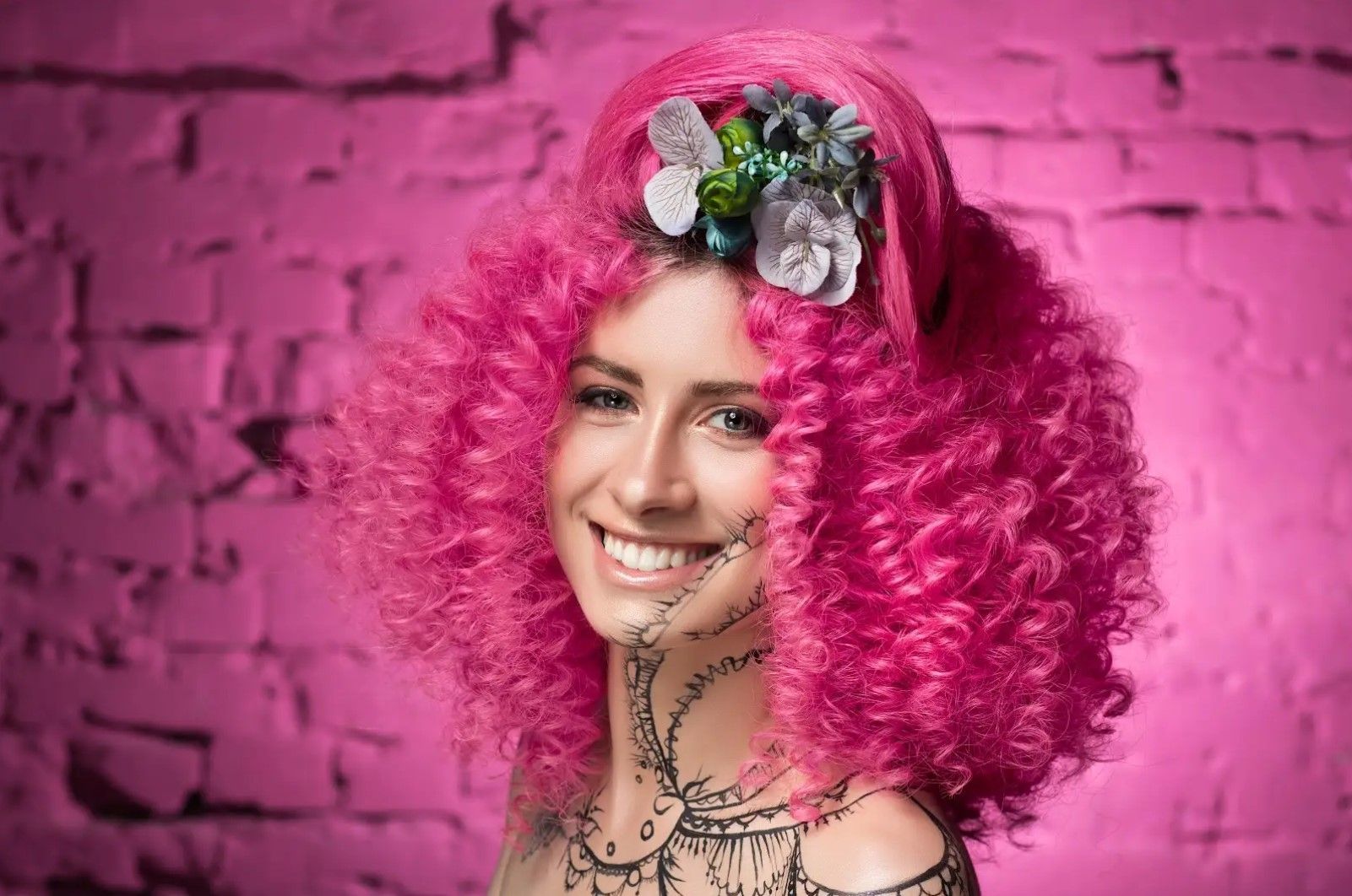 A woman with pink curly hair is smiling in front of a pink brick wall.