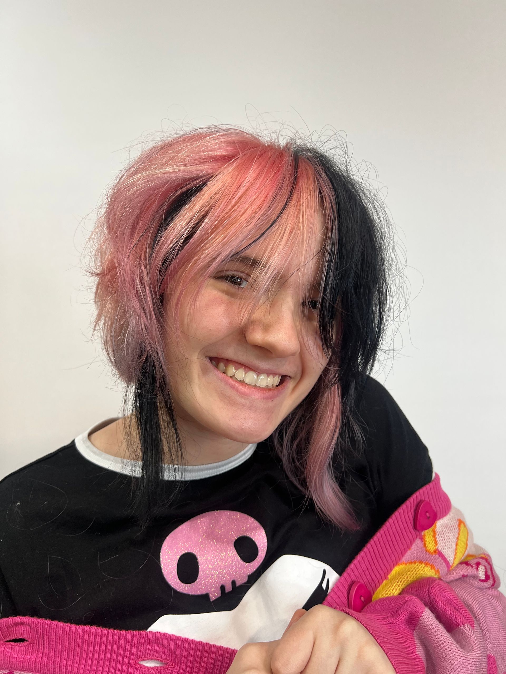 A girl with pink and black hair is smiling and wearing a pink sweater with a skull on it.